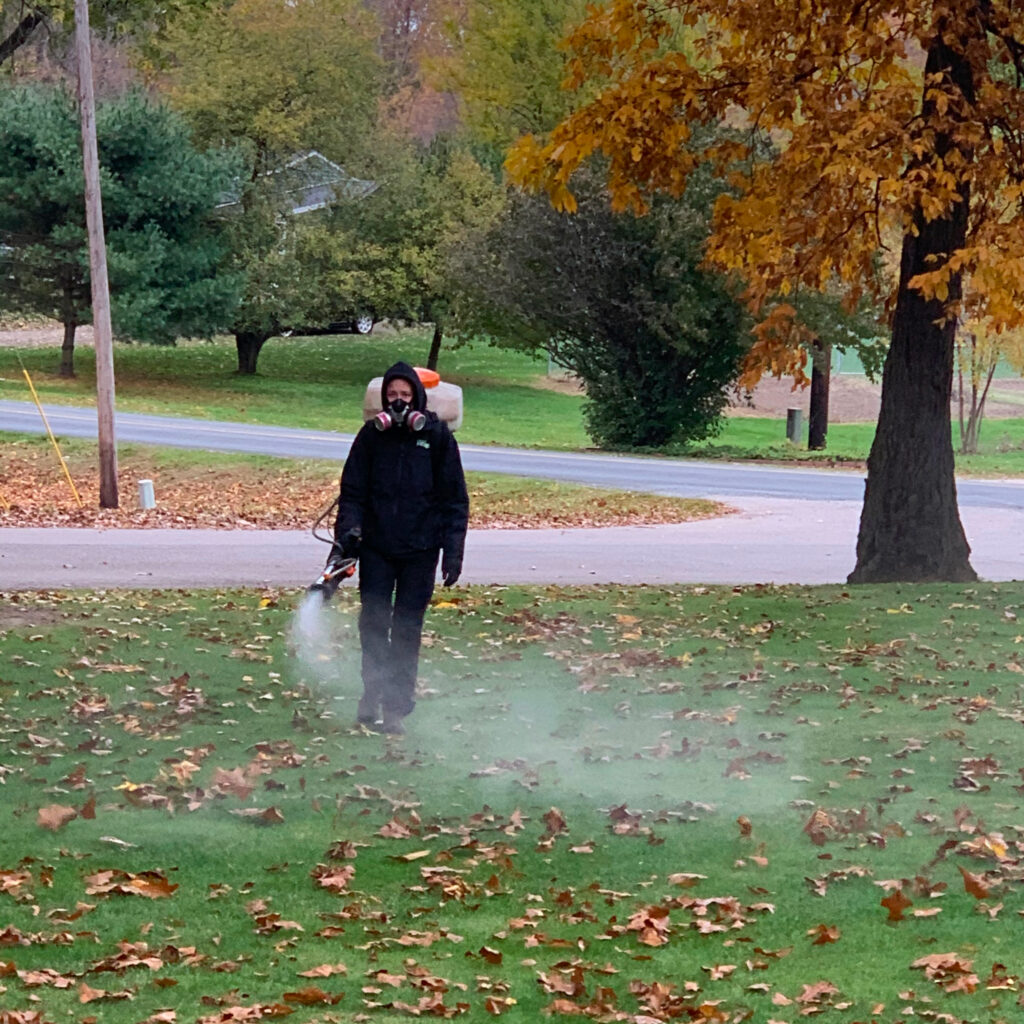 One of our Pest Pros technicians, dressed in protective gear, spraying pest control treatment across a lawn in the fall.